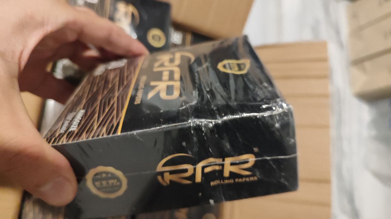 RFR Rolling Papers from Shang Go Technology Co. Ltd — a damaged black box with torn plastic wrapping and visible creases, held in a hand. The image shows the poor quality and disappointing experience with the product, with several similar boxes stacked in the background.