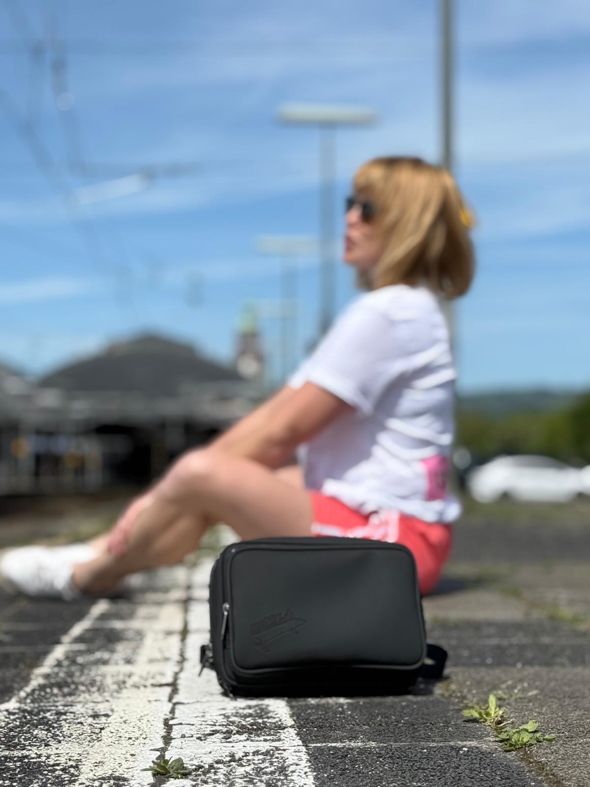 Eine Frau sitzt entspannt im Hintergrund auf einem Bahnsteig in der Sonne. Im Fokus im Vordergrund steht die schwarze HIGHTÄSCH Umhängetasche, die stabil auf dem Boden abgestellt ist und das dezente Logo zeigt